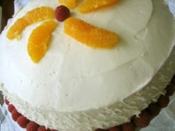 Cake covered in cream cheese frosting on a glass cake stand with raspberries surrounding the base of the cake and orange slices garnishing the top.