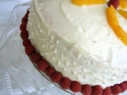 Side of a cake covered in cream cheese frosting on a glass cake stand with raspberries surrounding the base of the cake.