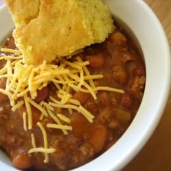 Chili in a white bowl with a piece of cornbread topped with shredded cheese.