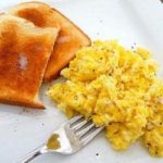 Scrambled eggs with 2 pieces of toast on a white plate with a fork.