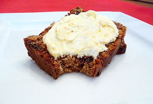 Bite out of a slice of date nut spice bread with cream cheese frosting on a white plate.