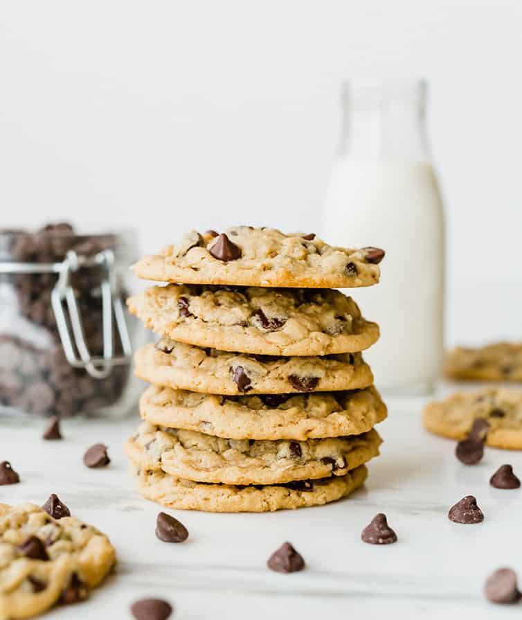 A stack of peanut butter-oatmeal chocolate chip cookies with chocolate chips and milk in the background.