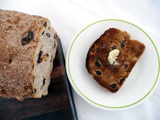 Slice of cinnamon raisin walnut bread on a white plate topped with a pat of butter.