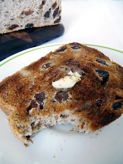 Slice of cinnamon raisin walnut bread topped with butter with a bite taken from it on a white plate.