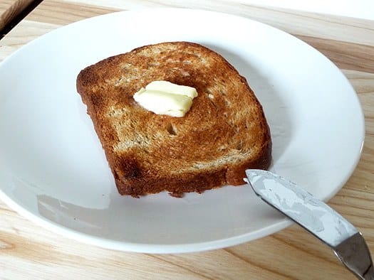 Slice of honey oatmeal sandwich bread on a white plate with a pat of butter on top.