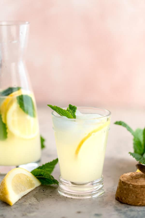 A glass of lemonade with mint and lemon slices, with a pitcher of lemonade in the background.
