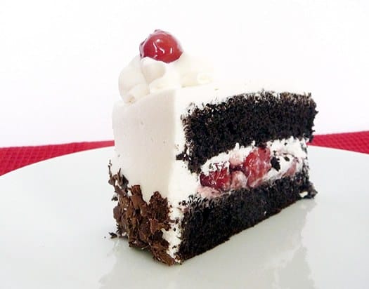 Side view of a slice of black forest layer cake topped with whipped cream and a cherry on a white plate.