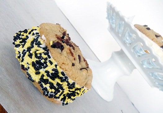 Chocolate chip cookie ice cream sandwich with vanilla ice cream rolled in black and gold sprinkles on a white dessert stand.