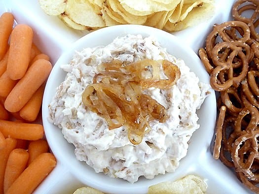 Overhead image of French onion dip topped with sautéed onions in a white bowl surrounded by pretzels, chips, and carrots.