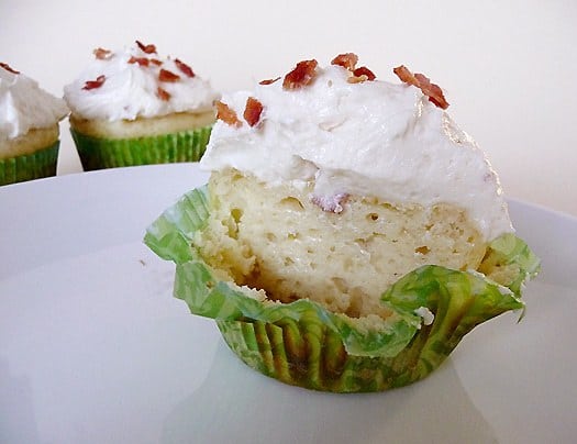 Pancake cupcake cut in half showing the texture topped with maple frosting and crumbled bacon on a white plate.