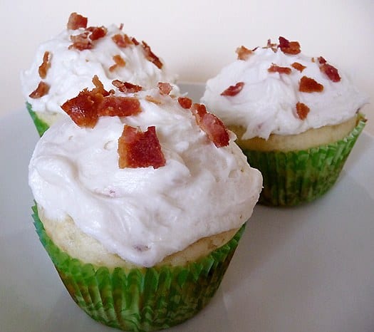 3 pancake cupcakes in green cupcake liners topped with maple frosting and sprinkled with crumbled bacon on a white plate.