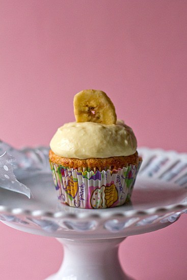 Banana cupcake topped with vanilla pastry cream and a banana chip on a white dessert stand.