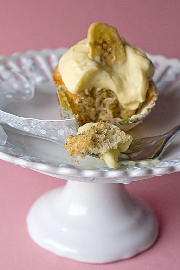 Banana cupcake topped with vanilla pastry cream and a banana chip on a white dessert stand with a fork taking a bite.