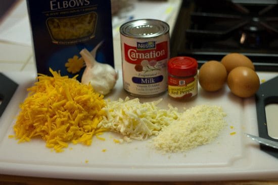 Ingredients for baked macaroni and cheese on a white cutting board including shredded cheese, noodles, garlic, milk, red pepper flakes, and eggs.
