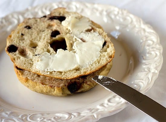 A blueberry bagel cut in half with cream cheese spread on top on a white plate with a knife.