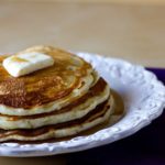 Stack of 3 pancakes topped with a pat of butter and maple syrup on a white plate.