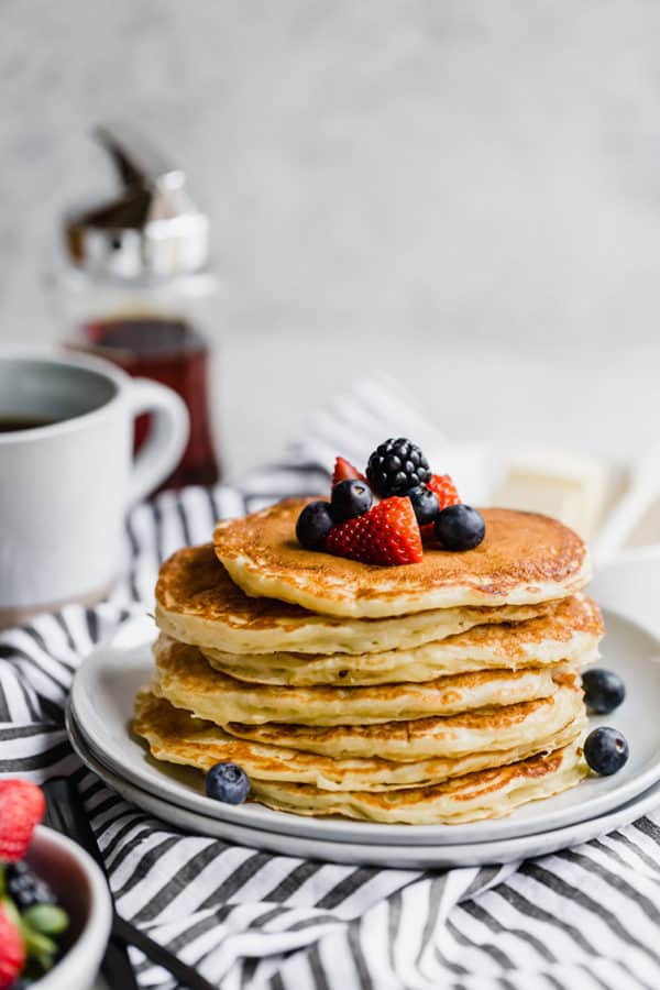 A stack of buttermilk pancakes with berries and a cup of coffee and syrup in the background.