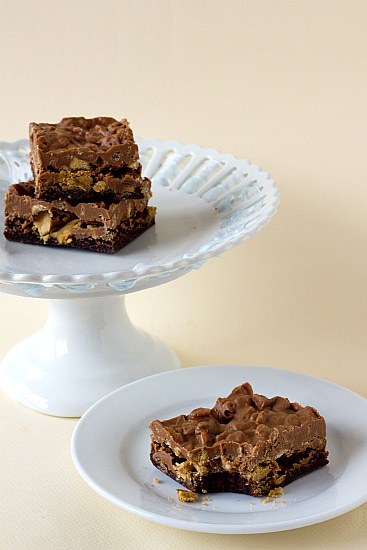 Peanut butter cup crunch brownie bars on a white and one with a bite taken from it on a white plate.
