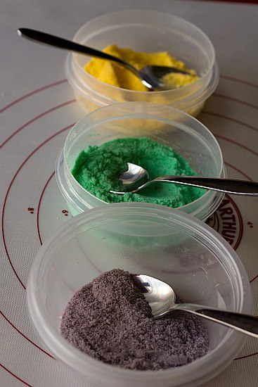 Yellow, green, and purple colored sugars in clear bowls with spoons.