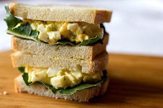 Stack of 2 egg salad sandwiches on a wood board.
