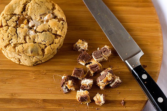 Overhead image of a stack of peanut butter Snickers cookies and chopped Snickers bars on a wood serving board with a knife.