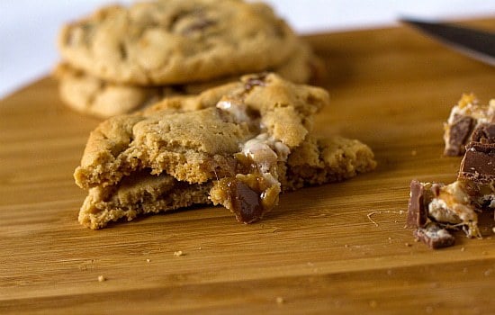 Peanut butter Snickers cookies on a wood serving board with one cookie broken in half showing the gooey texture.