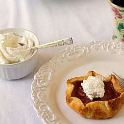 Rhubarb pie tartlet on a white plate with a fork and whipped cream in a white bowl with a spoon.