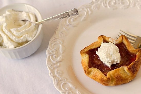 Rhubarb pie tartlet on a white plate with a fork and whipped cream in a white bowl with a spoon.