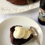 Overhead image of a slice of root beer float cake topped with vanilla ice cream on a white plate with a spoon.