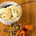 Scoops of dried apricot pistachio ice cream in a white bowl with a spoon with pistachios and dried apricots next to the bowl.
