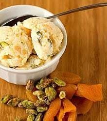 Scoops of dried apricot pistachio ice cream in a white bowl with a spoon with pistachios and dried apricots next to the bowl.