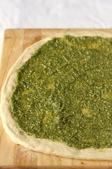 Pizza dough rolled out and topped with pesto on a wood cutting board before baking.