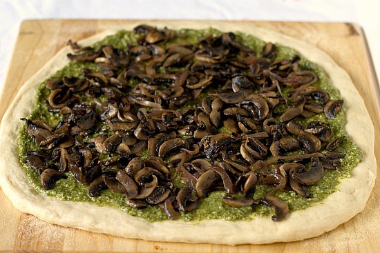 Pizza dough rolled out and topped with pesto and sautéed portobello mushrooms on a wood cutting board before baking.