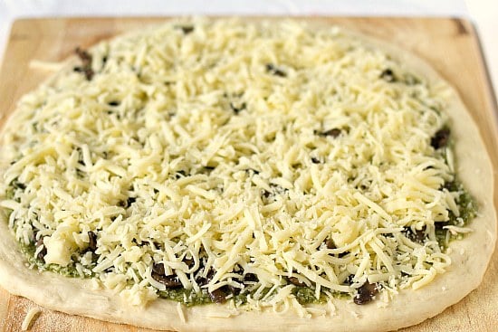 Pizza dough rolled out and topped with pesto, portobello mushrooms, and shredded cheese on a wood cutting board before baking.
