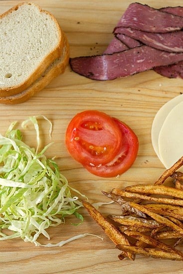 Ingredients for a homemade primanti brother's sandwich including pastrami, cheese, cabbage slaw, tomatoes, and french fries on a wood cutting board.
