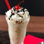 White Russian milkshake in a glass topped with whipped cream, chocolate sauce drizzle, and a cherry.