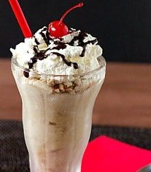 White Russian milkshake in a glass topped with whipped cream, chocolate sauce drizzle, and a cherry.