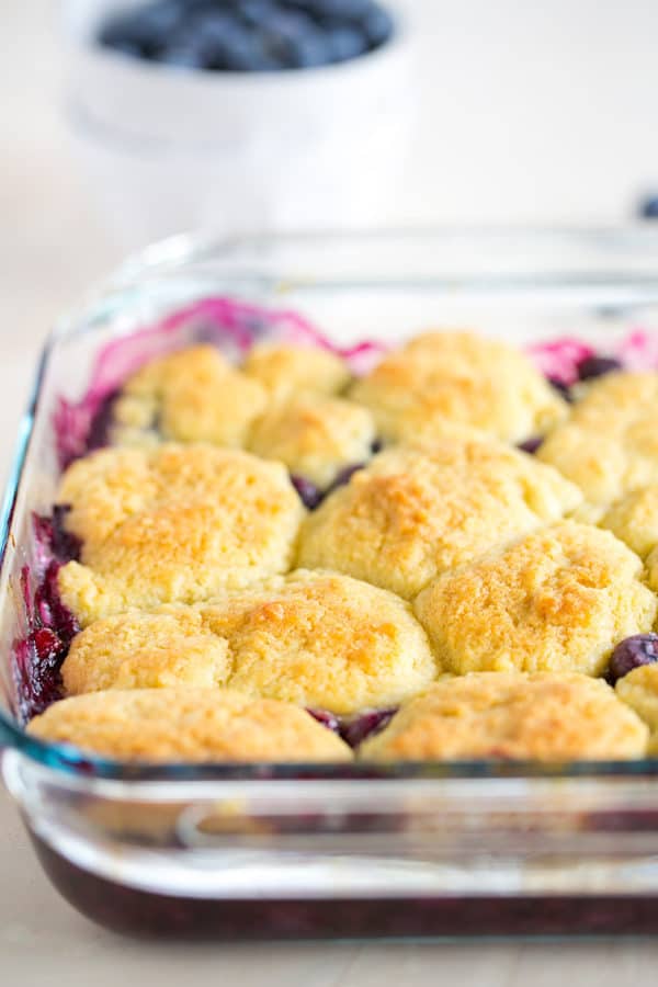 Baked blueberry cobbler in a glass pan with a bowl of blueberries in the background.