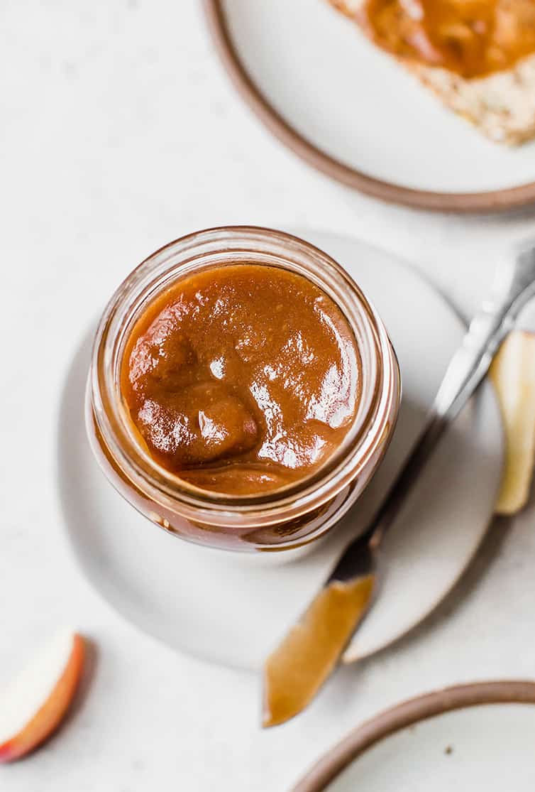 A jar of apple butter with a butter knife and piece of toast in the background.
