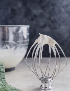 A whisk attachment standing upright with whipped cream on top, with a bowl of whipped cream behind.