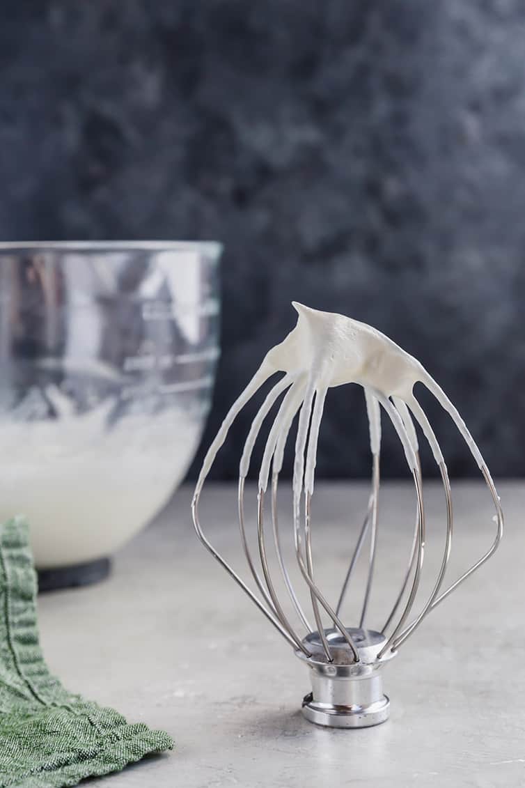 A whisk attachment standing upright with whipped cream on top, with a bowl of whipped cream behind.
