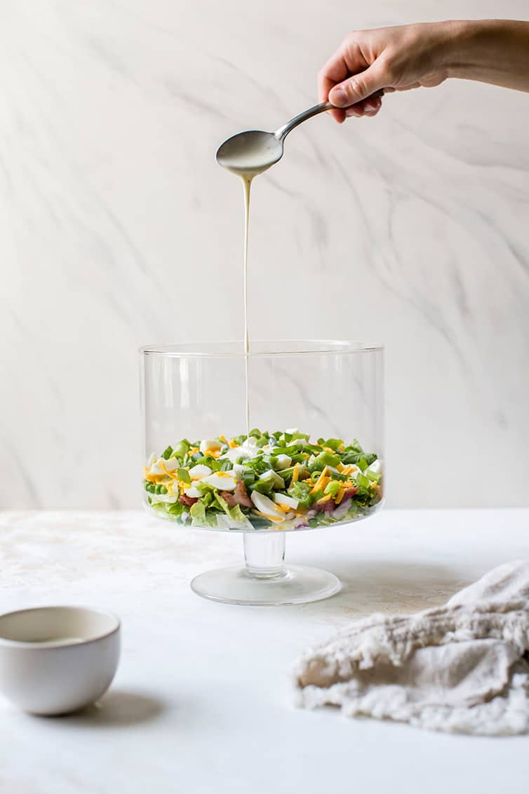 Drizzling the dressing over a layer of seven layer salad in a trifle bowl.