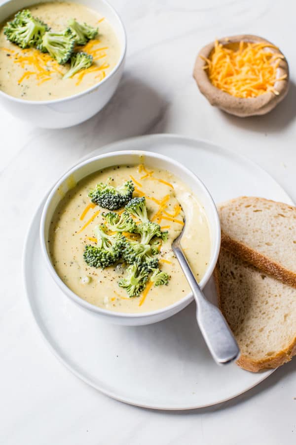 Two bowls of broccoli cheese soup with sliced bread and shredded cheese on the side.