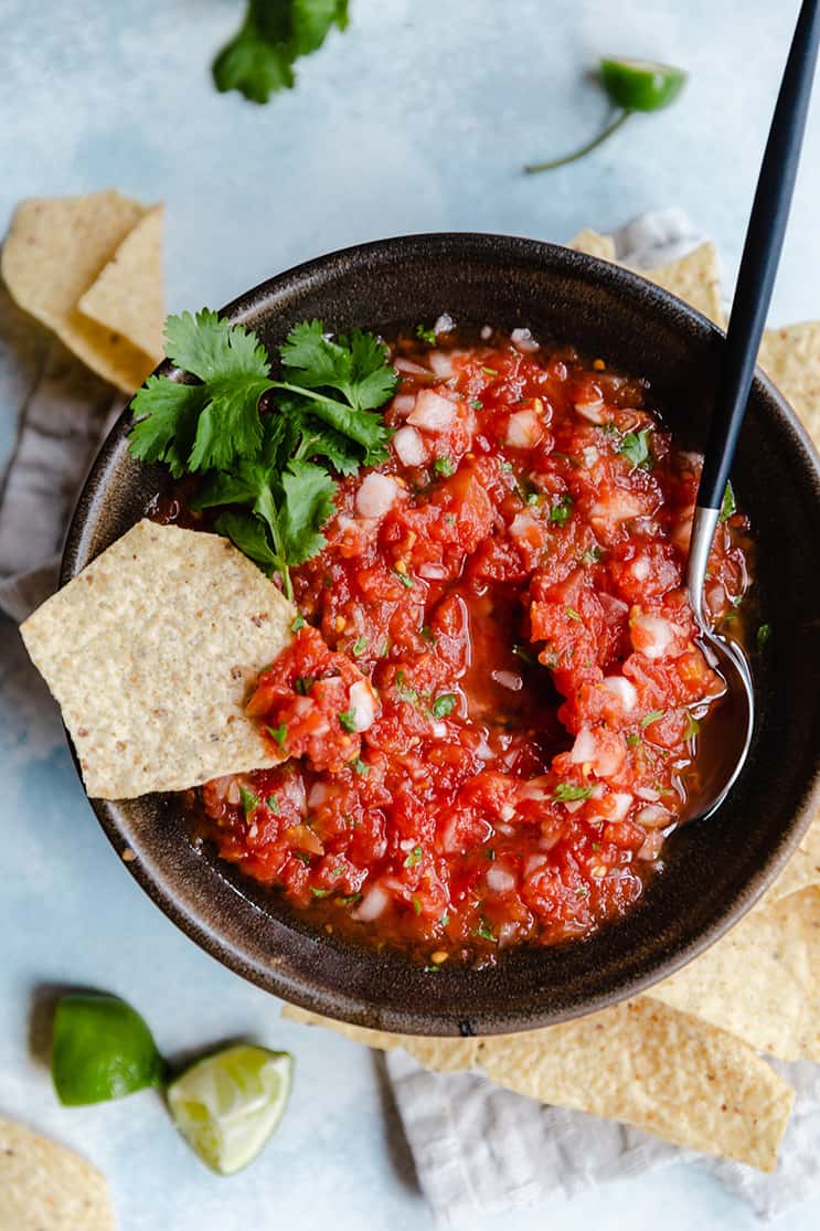 A bowl of salsa with a spoon on one side and a tortilla chip dipped in.