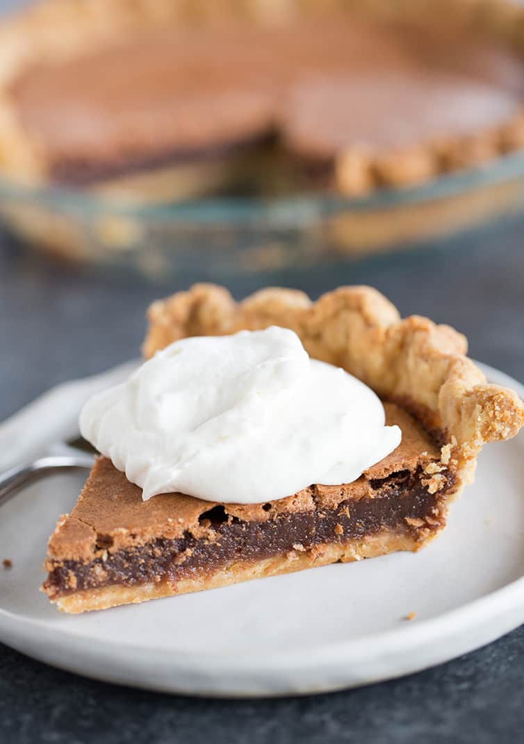 A slice of chocolate chess pie with whipped cream on top in front of the pie plate with the rest of the pie.