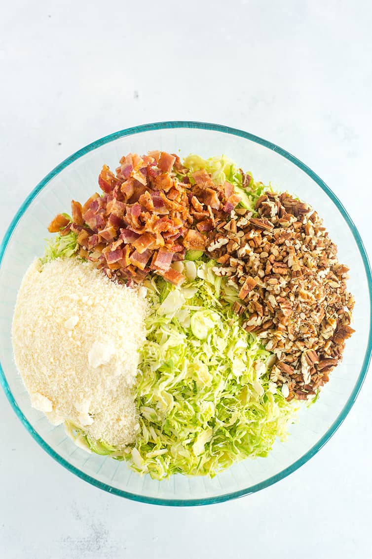 A bowl with shaved Brussels sprouts, chopped bacon, chopped pecans, and Parmesan cheese.