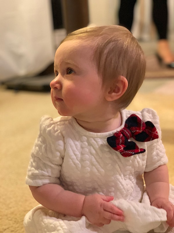 Baby girl sitting on the floor in a white dress with green and red bow.