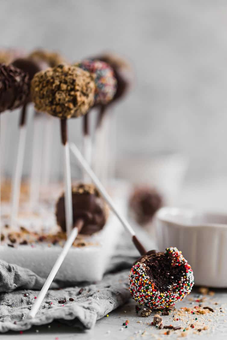 A chocolate cake pop with a bite taken out on the counter with its stick up and a block of chocolate cake pops behind it. 