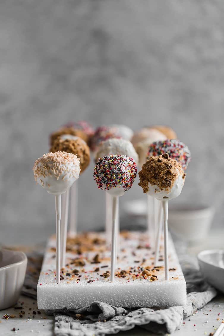 Vanilla cake pops standing up in a styrofoam block topped with sprinkles and cookie crumbs.
