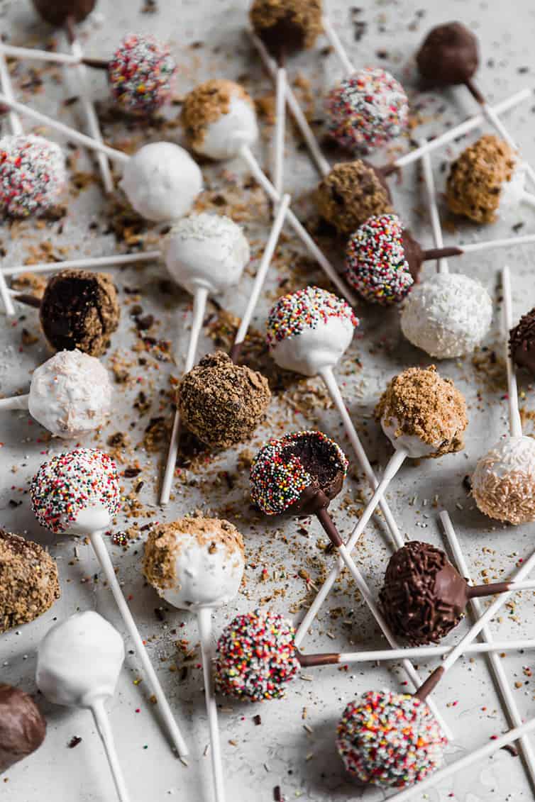 An overhead shot of tons of cake pops dipped in white and milk chocolate and coated with toppings laying on a surface.